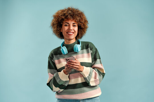 Smiling young woman with curly hair wearing headphones and a striped sweater while holding a smartphone against a blue studio background