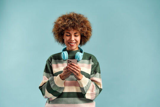 Smiling young woman with curly hair using a smartphone, wearing turquoise headphones and a striped sweater against a plain blue background.