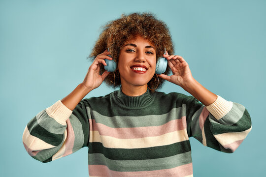 Smiling young woman with curly hair wearing headphones and a striped sweater, enjoying music against a bright teal background