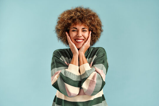 Beautiful young woman with curly hair holds her face in delight against a light blue studio background, wearing a cozy patterned sweater.