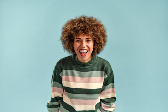 Joyful young woman with curly hair laughing in a colorful striped sweater against a clean turquoise background, centered portrait with space for text.