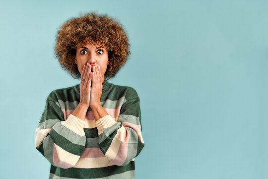 Surprised young woman with curly hair covering her mouth in shock, wearing a striped sweater against a plain light blue background with copy space.