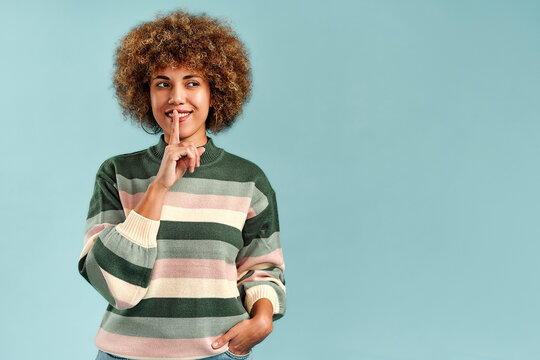Smiling young woman with curly hair wearing a striped sweater, making a quiet gesture with her finger to her lips against a blue background with copy space.