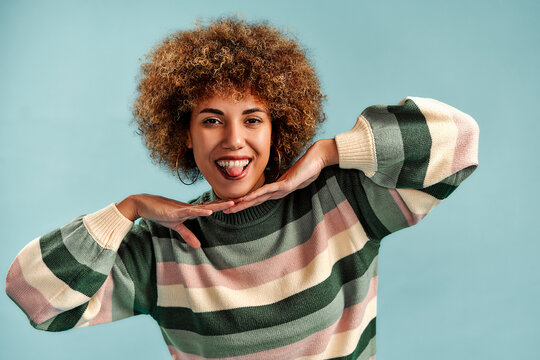 Smiling young woman with curly hair posing playfully with hands under her chin in a striped sweater against a light blue studio background.
