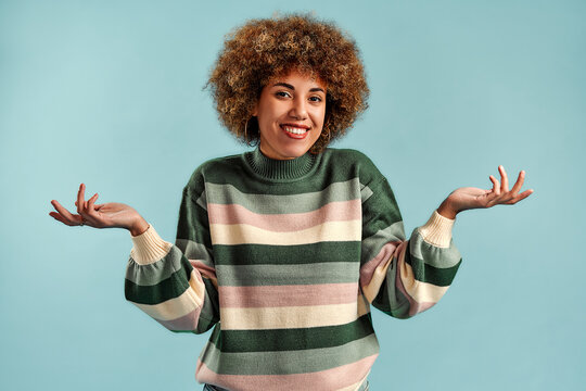 Smiling young woman with curly hair in a striped sweater shrugging with open hands against a light blue studio background.