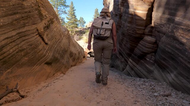 Female explorer wearing backpack walking through deep narrow sandstone slot canyon trail under bright sunlight during outdoor adventure in desert national park zion. Back view following of hiker