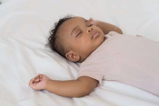 African American newborn adorable sleeping baby lying peacefully on white bedsheet with arms raised up, wearing soft pink bodysuit, curly dark hair, calm and serene infant nap time