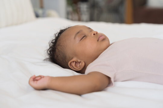 African American newborn adorable sleeping baby lying peacefully on white bedsheet with arms raised up, wearing soft pink bodysuit, curly dark hair, calm and serene infant nap time