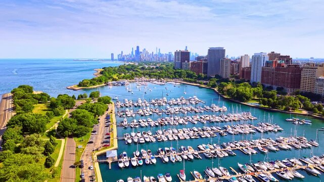 Moored yachts and sailboats in lakefront harbor, Chicago. Rows of motorboats and sailboats on floating piers, a parking lot, green trees, brick apartment buildings and the skyline.
