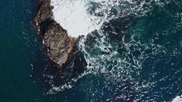 Foam from ocean waves moves around a large rock along the coast, seen from above during daylight.