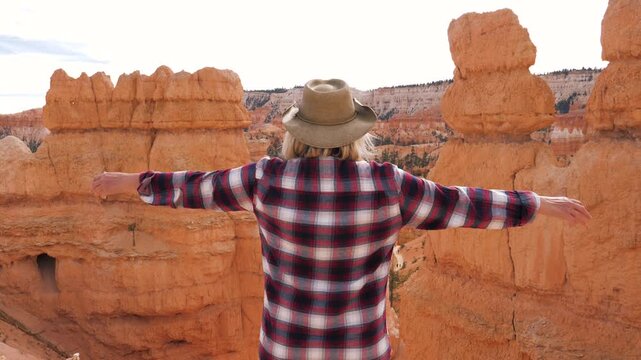 Sporty female hiker wearing hat standing on top cliff edge, raising her arms in triumph while looking at red orange rock hoodoos during mountain journey. Joy of successfully reaching the top trail