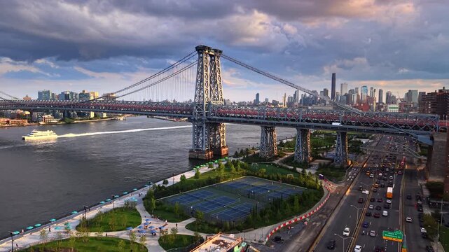 Steel bridge tower beside tennis courts and highway traffic, New York. Williamsburg Bridge cables, East River Park lawns, a ferry with wake and Brooklyn skyline under dusk clouds.