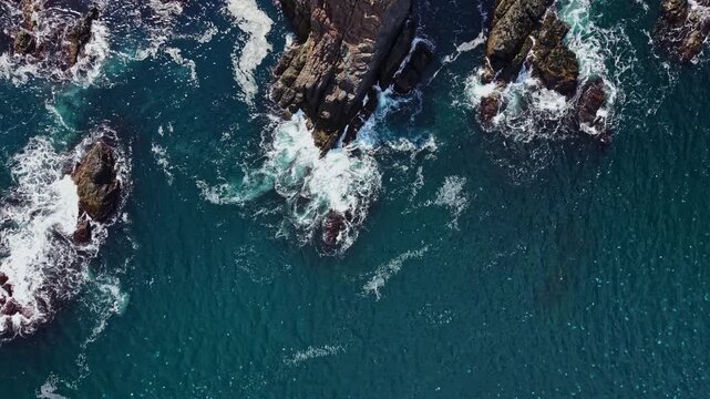 From an overhead view, ocean water moves around rocks along the coast, forming white foam where waves break against the shoreline during daylight.