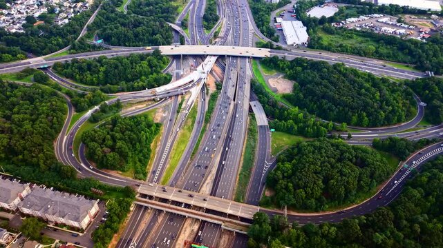 Highway interchange with curved ramps among forest, New Jersey. Multilane Garden State Parkway, overpasses, dense green trees, townhouses and warehouse lots with parked trailers.