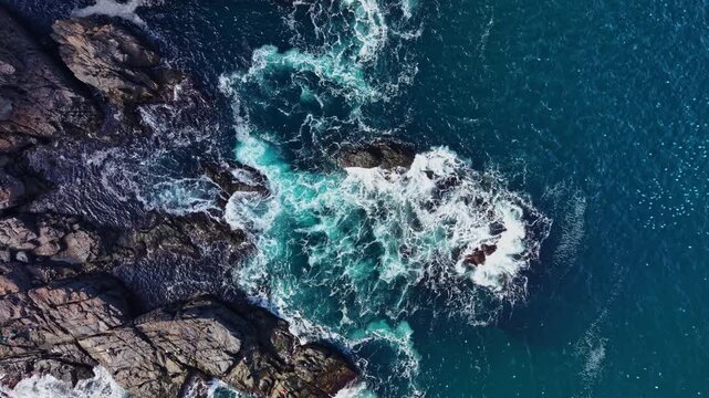 Waves move across blue ocean water and break into white foam against a rugged rock shoreline, seen from above during the day.