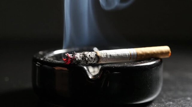 A close-up of a burning cigarette resting in a black ceramic ashtray with smoke against a dark background.
