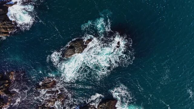 Water surges and forms foam as waves strike exposed rocks along the coast, seen from above during daylight.