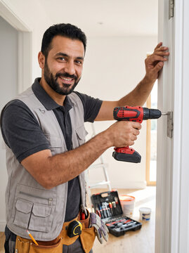 Smiling worker with cordless screwdriver fixing a door hinge in a bright home hallway, tool belt and open toolbox on the floor. Reliable handyman service and home repair concept