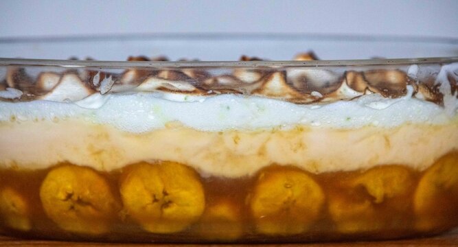 Chico balanceado, a traditional Brazilian layered dessert with caramelized bananas, vanilla custard and toasted meringue with lime zest, served in a glass baking dish on a wooden table 