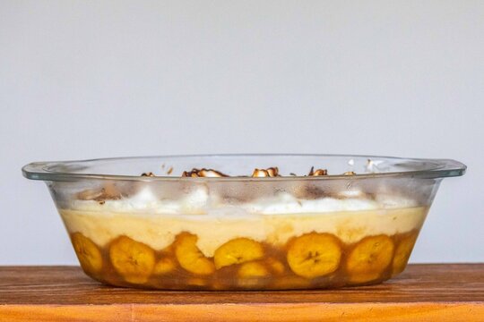 Chico balanceado, a traditional Brazilian layered dessert with caramelized bananas, vanilla custard and toasted meringue with lime zest, served in a glass baking dish on a wooden table 