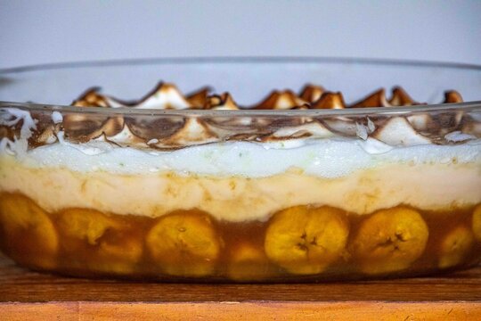 Chico balanceado, a traditional Brazilian layered dessert with caramelized bananas, vanilla custard and toasted meringue with lime zest, served in a glass baking dish on a wooden table 
