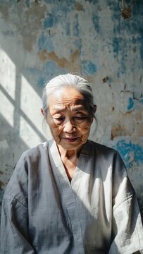 Portrait of an elderly Asian woman with gray hair, traditional clothing, and weathered skin, illuminated by natural light against a textured wall.