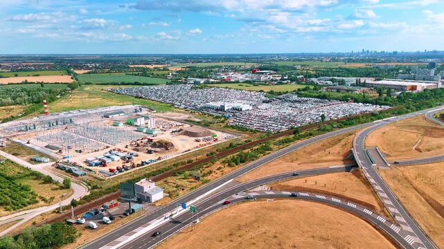 Construction borders car storage lots beside curving highway ramps. Highway ramps curve beside parked vehicles, railway tracks and an unfinished building.