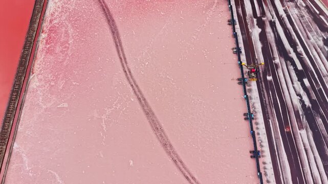 Salt extraction takes place in a large pond of pink water, with dark tracks crossing the surface and parallel roads and channels running along the edge.