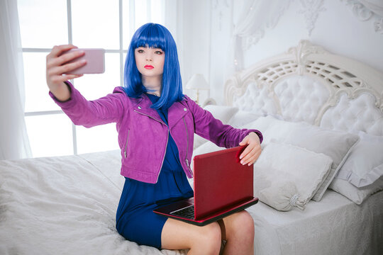 Fashion freak. Glamour emotionless synthetic girl, fake doll with blue hair is holding computer and doing selfie while sitting on the bed. Stylish beautiful woman in purple jacket in the bedroom. 