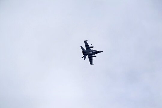Military fighter jet flying overhead in an overcast sky, viewed from below showing underside wings, twin engines and mounted equipment