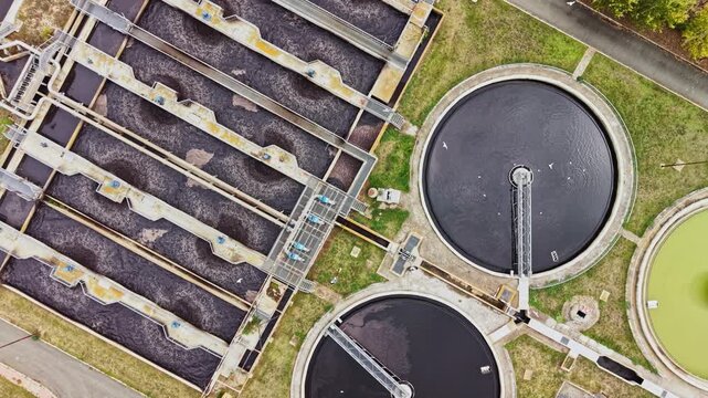 Seen from above during the day, wastewater flows through long treatment channels beside round clarifier tanks at a water treatment plant.