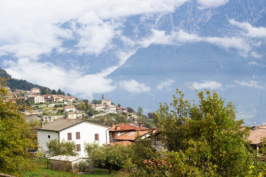 Mountain village with red tile rooftops, church tower, hillside homes, trees, and dramatic cloud covered alpine peaks in a scenic European landscape