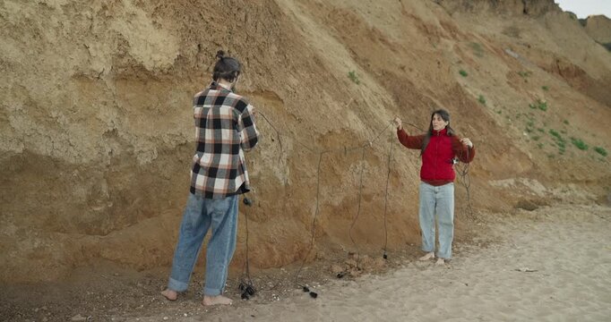 Two young adults on sandy beach beside eroded cliff untangling long cords in playful interaction while barefoot and casually dressed near shoreline with overcast daylight