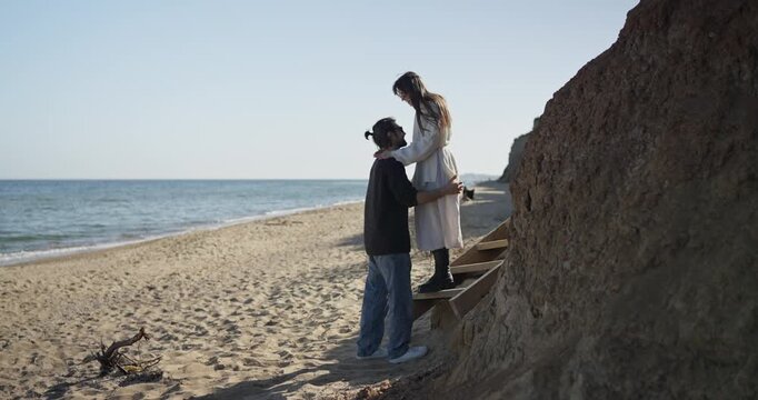 Two people walking hand in hand on sandy beach beside coastal cliff as man helps woman down wooden steps toward calm sea in bright daylight near waves and sky