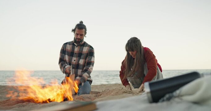 Two people tending campfire on sandy beach at dawn with warm flames and ocean horizon in background, one stacking wood while other arranges firepit and sand, soft morning light and calm coastal mood.