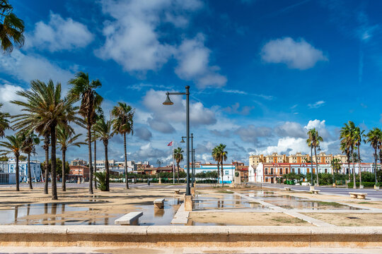 Square and promenade near Malvarrosa beach, Valencia, Spain, with tall palm trees, modern street lamps traditional colorful buildings under a blue cloudy sky.