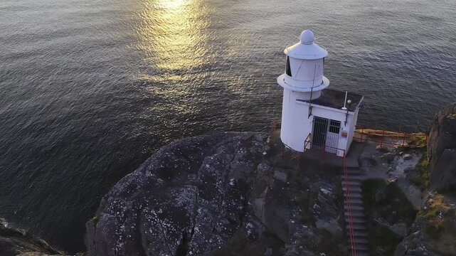 Aerial footage of white lighthouse perched on rugged rocky cliffs at edge of sea during sunset, with mountain silhouettes on distant horizon. Sheep's Head Lighthouse at sunset, County Cork, Ireland.