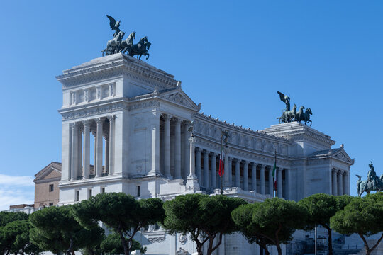 Vittoriano monument with bronze quadrigas and Italian flags under blue sky behind umbrella pines in Rome, Italy