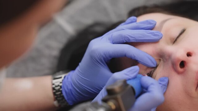 Close up of cosmetologist performing permanent makeup procedure on eyelid. Specialist using tattoo machine for microblading or cosmetic tattooing in a modern spa