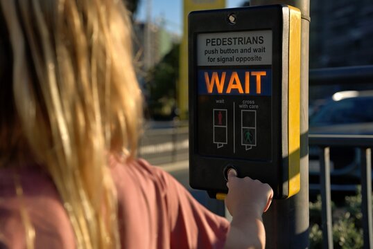 Girl pushes the wait button on a pedestrian crossing signal box by the roadside in daylight