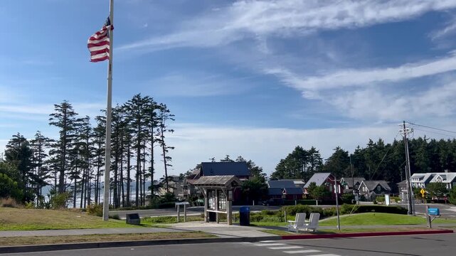 US flag whipping in a stiff ocean breeze above the community green of Seabrook, Washington, overlooking shingled homes, tall pines and a kiosk.