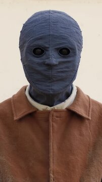 Charcoal cloth masked figure giving a slow nod against cream