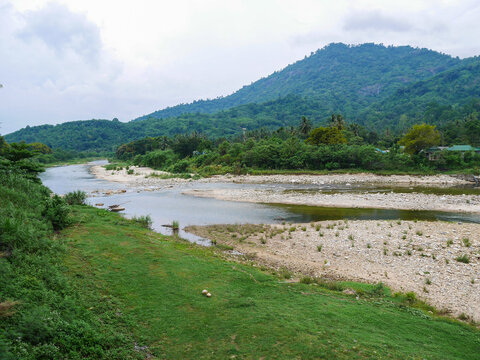 Scenic landscape of a wide clear river with a gravel sandbar, flowing through lush green grass, tropical trees, and a mountain range under an overcast sky.