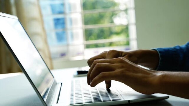 Hands typing on laptop at home workspace, Close-up of hands typing on a laptop at a bright desk near a window, showing remote work, online study, and modern digital communication.