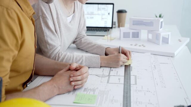 Architects collaborating on building blueprints, discussing design details and making notes with a ruler and pen, working on a new project in a modern office