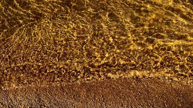 Sunlit shallow water glides gently over coarse golden sand at beach edge. Clear ripples cast dancing caustic patterns across lake floor, providing serene natural view full of warm calm ambiance.