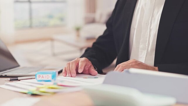 Businessman reviews financial invoice paperwork beside paid stamp laptop on office desk. Corporate tax billing management, expense settlement audit, accounting payment processing workflow.
