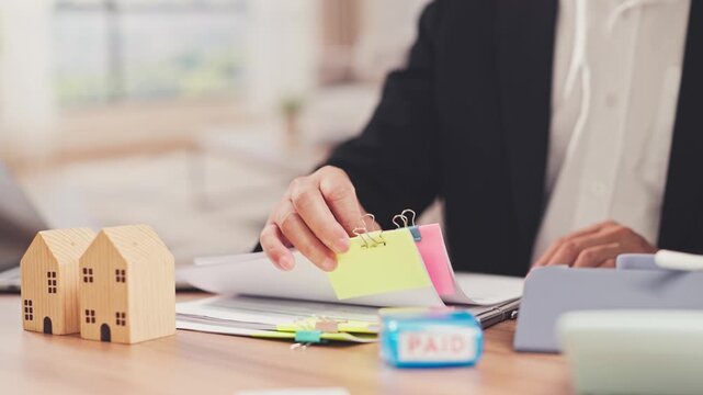 Wooden miniature house models stand beside paperwork while person writes in background. Real estate mortgage loan calculation, property investment analysis, residential property appraisal.