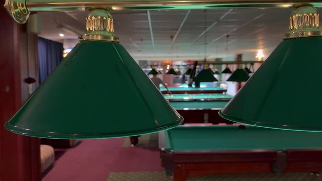 Traditional green pendant lights illuminate tables, people play inside cozy billiard club. Classic cue sport hall atmosphere, blurred background with players during tournament.