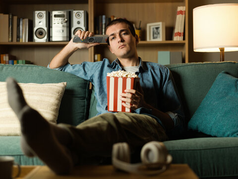 Bored young man holding TV remote control and popcorn box on couch at home
